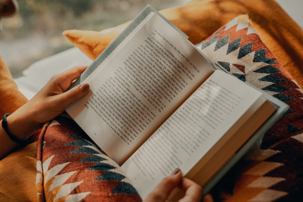 An opened book is held in the hands of the reader and perched on aztec printed orange, blue and cream cushions. Arts & humanities and social sciences reader.
